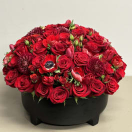 Low dome of red roses and mixed red blooms arranged in a round black bowl.