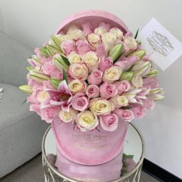 Pink and cream roses with pink lilies arranged in a round pink hat box with a card.