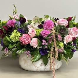 Low mixed arrangement of pink roses, purple blooms, and green accents in a white marble bowl