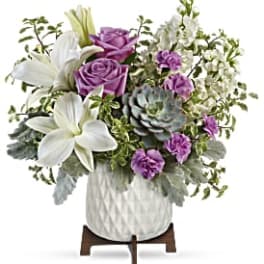 Arrangement of white lilies, lavender roses, purple carnations and a succulent in a white pot on wooden stand