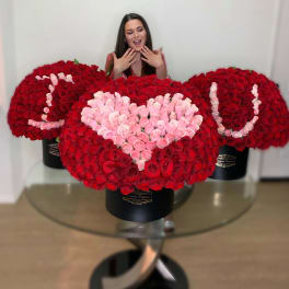 Three large red and pink rose hatbox arrangements spelling I heart U on a glass table