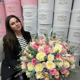 Large pink hatbox arrangement of pastel roses with orchids, next to a seated woman.