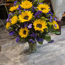 Bouquet of yellow sunflowers and purple filler flowers in a clear glass vase.
