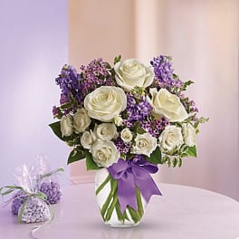 White roses and purple flowers arranged in a glass vase with a purple ribbon
