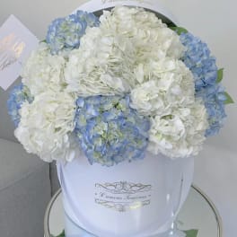 Round box of white and light blue hydrangeas with a card on a mirrored table
