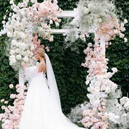Bride in a white gown beside a floral wedding arch with white and blush flowers