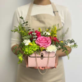 Person in an apron holding a pink purse-shaped box filled with pink, white, and green flowers.
