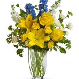Yellow lilies and roses with blue and white flowers in a clear vase