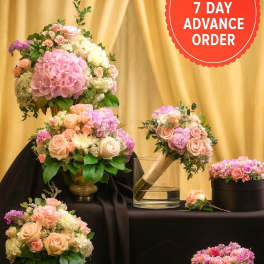 Set of blush and pink rose arrangements with hydrangeas in gold vases, boxes, and a bouquet on dark draped fabric