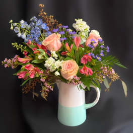 Mixed bouquet of pastel roses, blue delphinium, and purple daisies in a two-tone pitcher