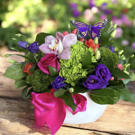 Colorful flower arrangement in a white vase with a pink ribbon and butterfly accent