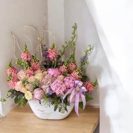 Pink and cream floral arrangement in a white bowl vase with a lavender ribbon