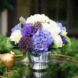 Purple and white flower arrangement in a silver vase beside a small gold bird figurine