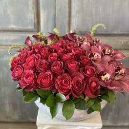 A low arrangement of red roses and burgundy orchids in a white bowl.