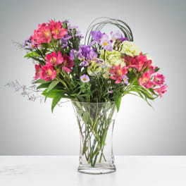Tall mixed bouquet of pink alstroemeria, purple blooms, and green carnations in a clear glass vase