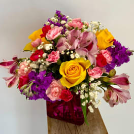 Bright mixed bouquet with yellow roses, pink carnations, and purple blooms in a pink cube vase