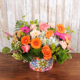 Low arrangement of orange and pink roses and gerbera daisies with blue hydrangea in a colorful round container