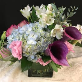 Low arrangement with blue hydrangea, purple calla lilies and pink blooms in a glass cube vase