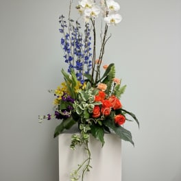Tall floral arrangement with white orchids, blue delphinium, and orange roses