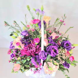 Basket arrangement of purple, pink, and peach flowers with a white handle