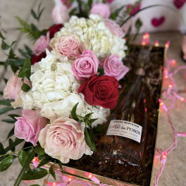 Pink and red roses with white hydrangeas in a gift box beside a wine bottle