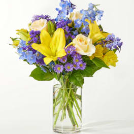 Bouquet of yellow lilies, cream roses, and purple flowers in a clear glass vase