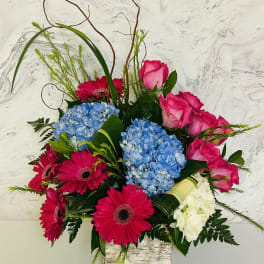 Bright bouquet of pink roses, blue hydrangeas, and magenta gerbera daisies in a square vase