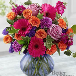 Bright bouquet of pink gerbera daisies, orange roses, and purple blooms in a round blue glass vase