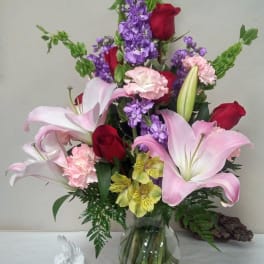 Mixed bouquet of pink lilies, red roses, purple flowers, and carnations in a glass vase