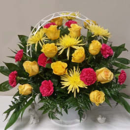 Yellow roses, pink carnations, and yellow spider mums in a white basket arrangement