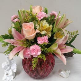 Pink lilies and roses arranged in a red glass vase with a small angel figurine
