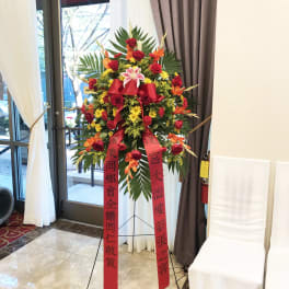 Tall standing floral spray with red ribbons and mixed red, yellow, and orange flowers