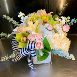 White and pink floral arrangement in a square vase with a striped ribbon