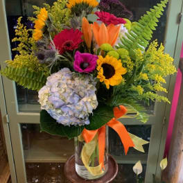 Tall mixed arrangement of sunflowers, lilies, gerbera daisies, hydrangea in a glass vase with orange ribbon