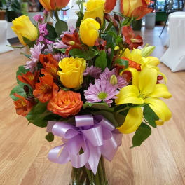 Mixed bouquet of yellow and orange roses with lilies and purple daisies in a glass vase