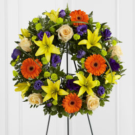 Circular floral wreath on a stand with yellow lilies, orange gerberas, cream roses, and purple blooms