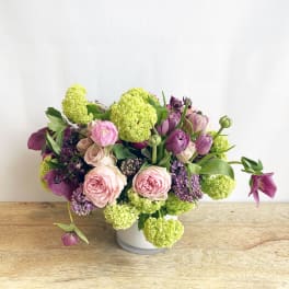 Low arrangement of pink roses, purple blooms, and green flower clusters in a white vase