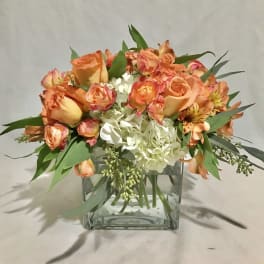 Short arrangement of orange roses and white hydrangeas in a clear glass cube vase