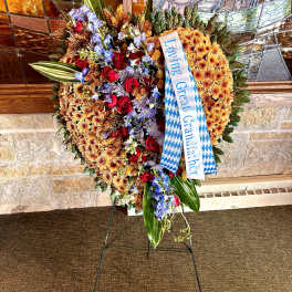 Heart-shaped standing funeral spray of orange mums with red roses, blue flowers, ribbon, and large green leaves on an easel