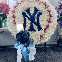 Large round standing wreath of white flowers with red accents and a blue floral emblem, finished with a dark bow.