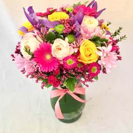 Bright mixed bouquet of pink, yellow, purple, and white flowers in a clear glass vase with a pink ribbon.