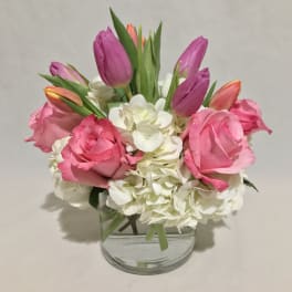 Compact arrangement of pink roses, purple tulips, and white hydrangeas in a clear glass vase