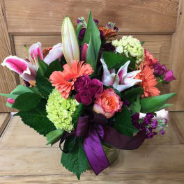 Mixed bouquet of lilies, roses, and gerbera daisies in a glass vase with a purple ribbon