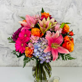 Mixed bouquet of pink lilies, orange roses, and purple flowers in a glass vase