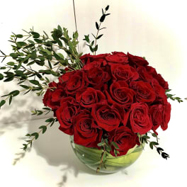 Round arrangement of red roses in a clear glass bowl vase with trailing greenery accents.