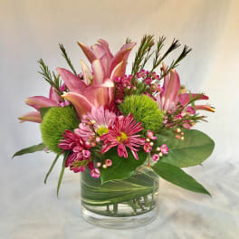 Compact arrangement of pink lilies and daisies with green pom blooms in a leaf-wrapped glass vase
