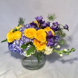 Short glass vase of yellow roses, blue hydrangeas, and purple flowers in a compact mixed arrangement