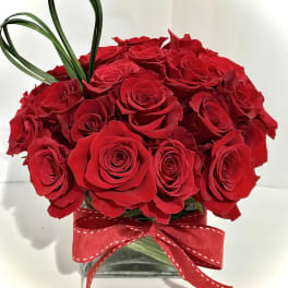 Compact arrangement of red roses in a glass cube vase with red ribbon and looped grass accent