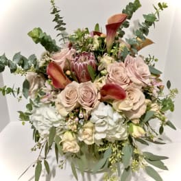 Round arrangement of blush roses, white hydrangeas, and orange calla lilies in a clear glass vase