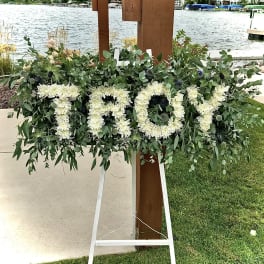 Standing tribute spray spelling TROY in white flowers with lush greenery on an easel